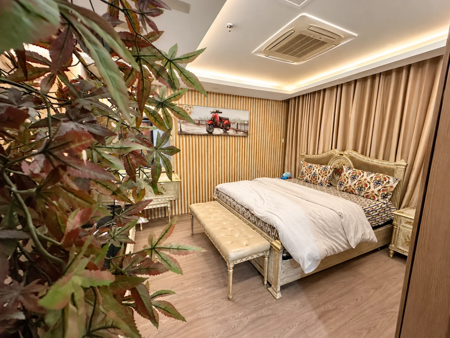 Bedroom with a gold-patterned headboard, bench and decorative indoor foliage