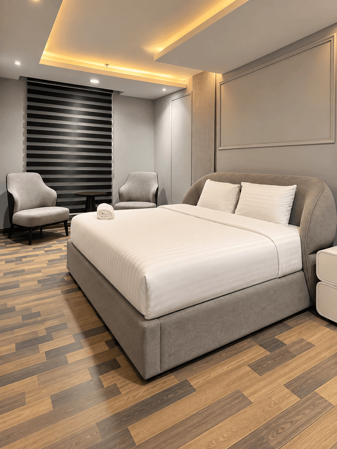 Grey upholstered bed with white linen, bedside tables and a seating corner beneath warm ceiling lighting