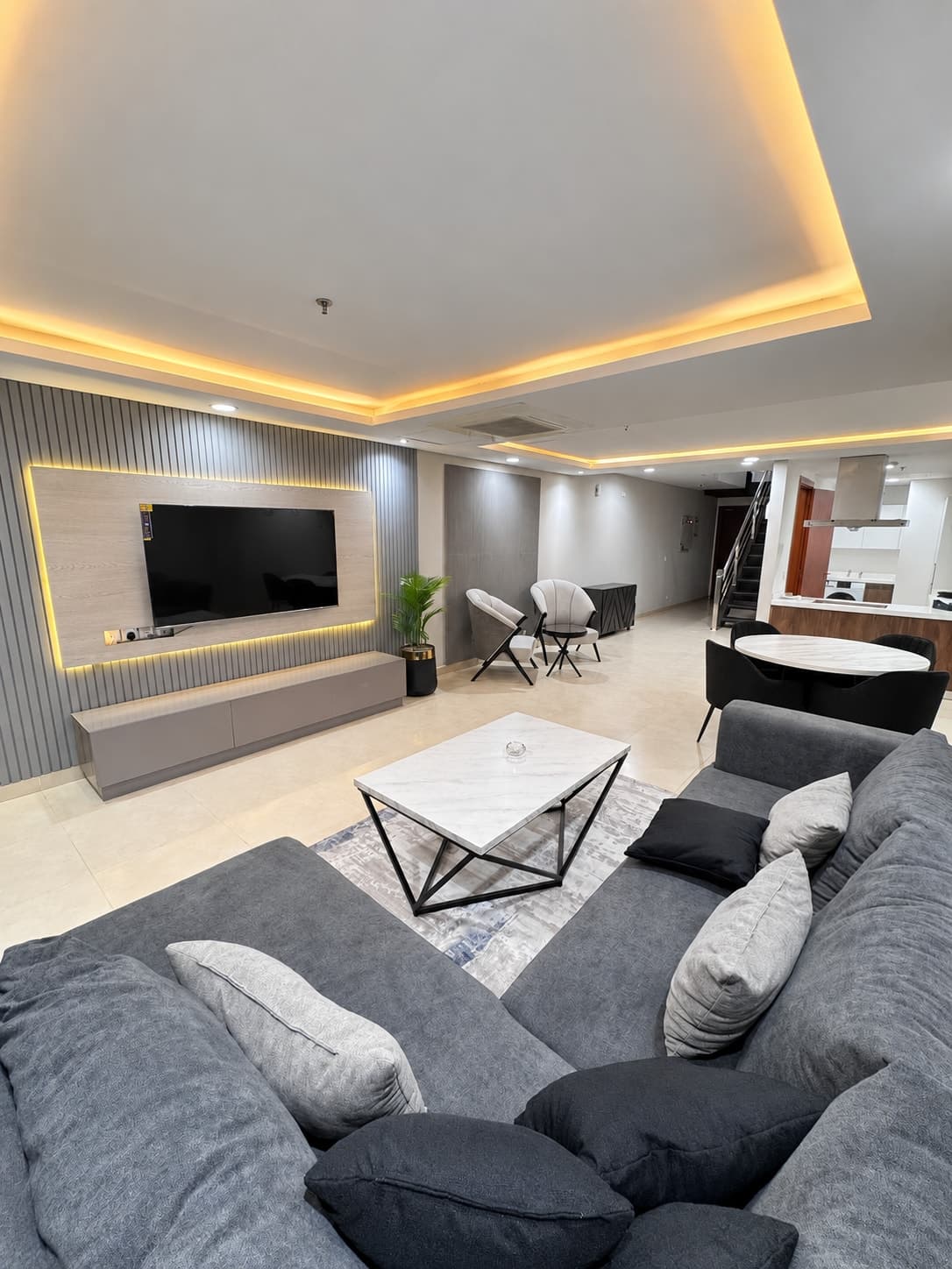 The Penthouse 2 lounge with a charcoal sectional sofa, pale coffee table and backlit TV wall, opening to the dining area and stairs