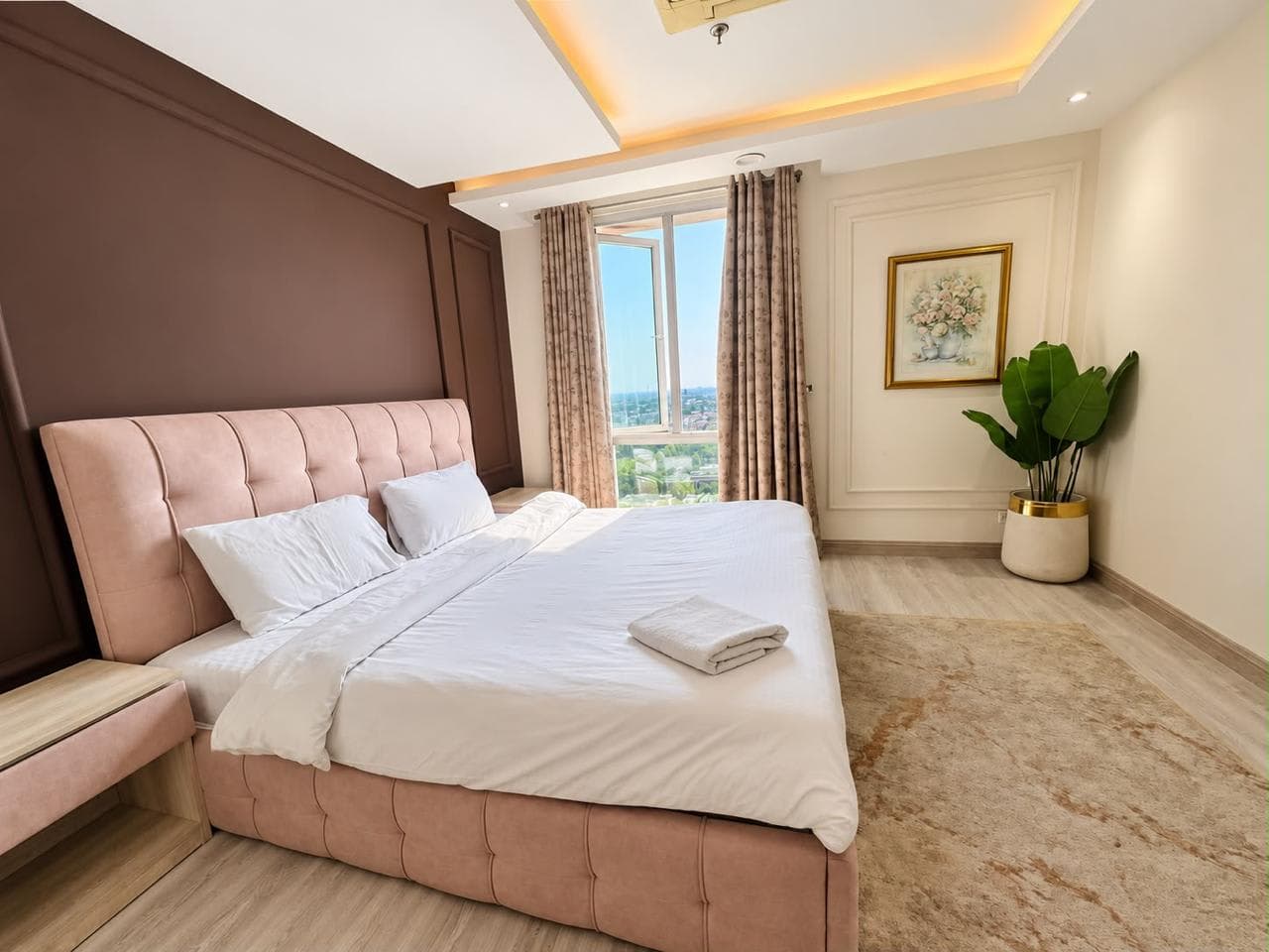 Blush upholstered bed beside a window, with a soft rug, framed floral artwork and a tall plant
